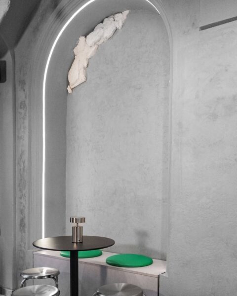 Nowoczesne wnętrze kawiarni o minimalistycznym charakterze – betonowe ściany z surową fakturą, delikatne podświetlenie LED w łuku oraz metalowy stolik z zielonymi siedziskami. Przestrzeń łączy industrialny chłód z nowoczesną estetyką.