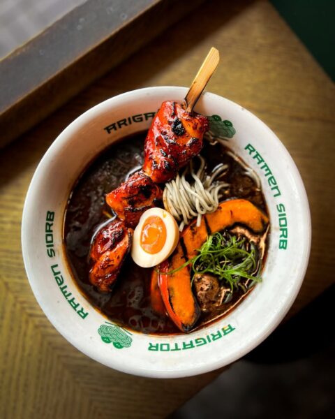 Duże pudełko wyłożone papierem pergaminowym, wypełnione starannie ułożonymi porcjami świeżych, surowych makaronów ramen, w spiralnych pęczkach, na drewnianym blacie kuchennym.