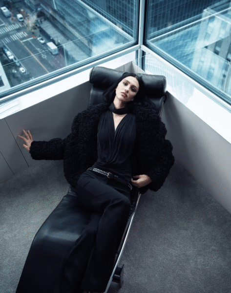 Kobieta w czarnym futrzanym płaszczu i eleganckim topie z dekoltem typu halter, leżąca na designerskim szezlongu przy panoramicznym oknie biurowca. Stylizacja utrzymana w klimacie miejskiego minimalizmu z nutą mocy.