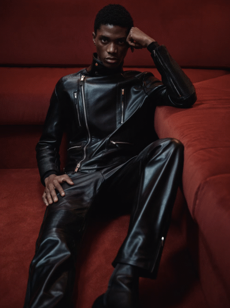 Mężczyzna w dopasowanym czarnym kombinezonie ze skóry z wieloma suwakami i przeszyciami, siedzący na czerwonej sofie. Ujęcie podkreśla kontrast tekstur i siłę sylwetki w estetyce rockowego luksusu.
