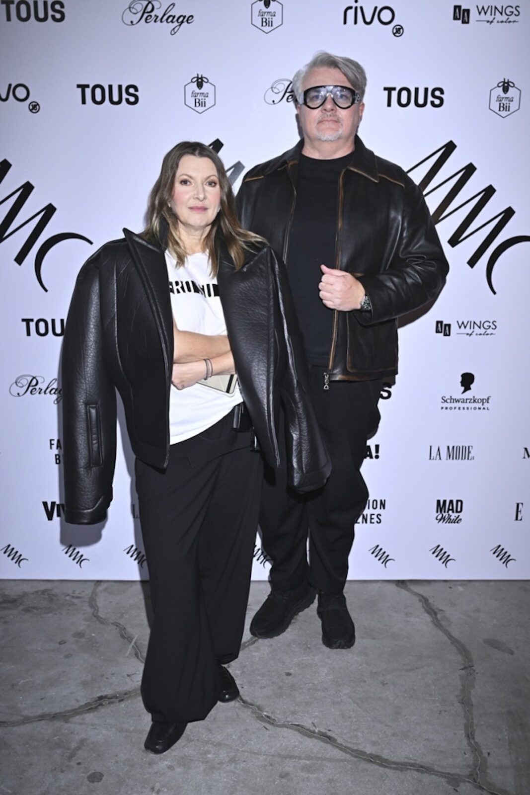 Zdjęcie z premiery kolekcji MMC w Warszawie – para w czarnych stylizacjach utrzymanych w minimalistycznym, nowoczesnym tonie. Kobieta ma na sobie oversize’ową kurtkę skórzaną zarzuconą na ramiona, białą koszulkę z nadrukiem i szerokie czarne spodnie, natomiast mężczyzna nosi czarną kurtkę skórzaną, T-shirt i spodnie w tym samym kolorze. Oboje pozują na tle ścianki z logotypami marek partnerskich, w tym Tous, Perlage i Schwarzkopf, w surowym, industrialnym otoczeniu betonu i reflektorów eventowych.