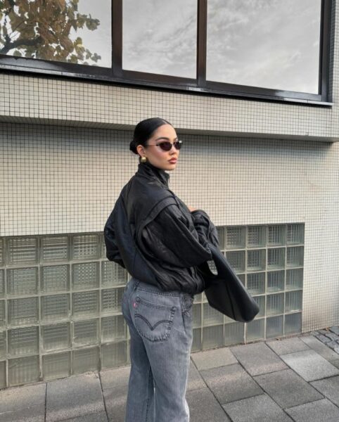 Kobieta w stylizacji "vintage cool" na zewnątrz. Ma na sobie ponadczasową czarną kurtkę typu bomber (lub luźną skórzaną/skóropodobną kurtkę) i klasyczne, szare dżinsy o prostym kroju (Levi's). Uzupełnieniem są modne, lecz proste, owalne okulary przeciwsłoneczne i złote kolczyki koła. Włosy są elegancko upięte w gładki kok, a cała stylizacja łączy luz z elegancją, co jest cechą modowych klasyków.