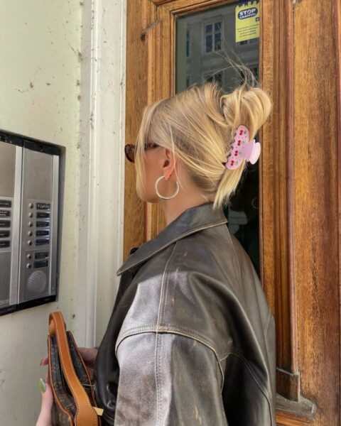 Kadr od tyłu przedstawia jasnowłosą kobietę z upiętymi włosami za pomocą różowej klamry z wiśniami, co jest przykładem szybkiej fryzury dla leniwych. Kobieta stoi przodem do domofonu w ciemnej, skórzanej kurtce, trzymając torebkę z monogramem.