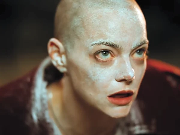 Kobieta z ogoloną głową i twarzą pokrytą białą, matową substancją, spoglądająca intensywnie w bok. Kadr o ekspresyjnym oświetleniu, ukazujący fizyczną i emocjonalną przemianę bohaterki w filmie Bugonia.