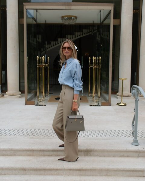 Kobieta o blond włosach, ubrana w błękitną koszulę i szerokie, beżowe spodnie, stoi na kamiennych schodach przed luksusowym budynkiem ze szklanymi drzwiami i marmurowymi kolumnami. Trzyma w ręku szarobeżową torebkę. Ma na sobie okulary przeciwsłoneczne w jasnej oprawie.