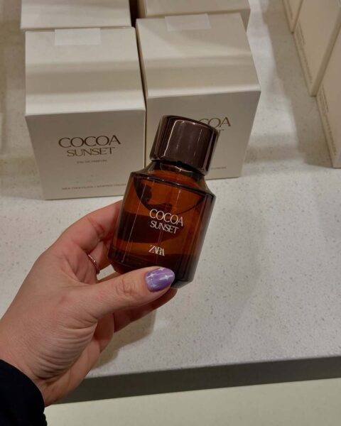 Ręka trzyma bursztynowy flakon perfum Zara Cocoa Sunset (Eau de Parfum) z brązową zakrętką. Flakon jest na tle białych pudełek z napisem "COCOA SUNSET | MILK CHOCOLATE | WHIPPED CREAM". To popularne i niedrogie czekoladowe perfumy na jesień 2025.