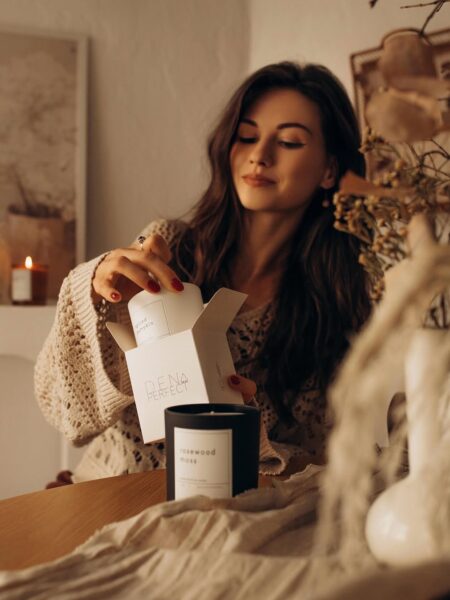 Urocza młoda kobieta o długich, brązowych włosach, ubrana w przytulny, beżowy sweter z dzianiny, siedzi w ciepło oświetlonym, jesiennym wnętrzu. Delikatnie otwiera białe opakowanie z napisem "DEN PERFECT", które skrywa małą, białą świecę zapachową. Obok leży większa, czarna świeca z etykietą "rosewood moss". Cała scena tworzy przytulną, jesienną atmosferę i przywodzi na myśl dom pachnący jak butik w Paryżu lub luksusowe SPA. To idealna chwila na hygge z ekskluzywnymi zapachami i relaks.