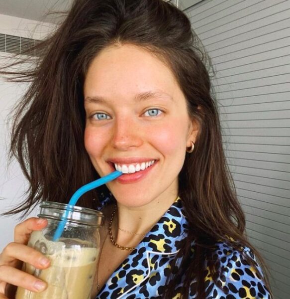 Modelka Emily DiDonato uśmiecha się szeroko, pijąc mrożoną kawę przez niebieską słomkę, a jej piękny, szeroki uśmiech z idealnie białymi zębami jest centralnym punktem kadru. Jej promienny wygląd przypomina o konieczności dbania o zdrowie jamy ustnej, zwłaszcza w obliczu potencjalnych problemów, takich jak ozempicowe zęby.