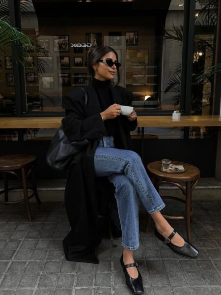 Kobieta siedzi przy stoliku w kawiarni na zewnątrz. Jest ubrana w długi, czarny płaszcz z poduszkami na ramionach, czarny golf, dżinsy o prostej nogawce oraz czarne buty z paskiem (mary jane). Pije z białej filiżanki i ma na sobie duże, ciemne okulary przeciwsłoneczne. Na stoliku obok widać szklankę wody.