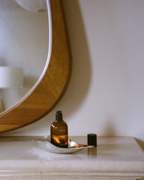 Butelka kosmetyku marki Aesop na marmurowej komodzie. Obok widoczne jest duże, drewniane lustro o nieregularnym kształcie.