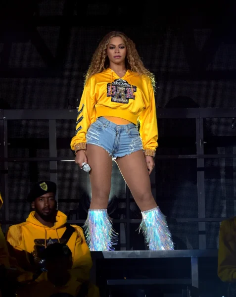 Beyoncé podczas występu na festiwalu Coachella – artystka w żółtej bluzie z logo „BAK”, krótkich jeansowych szortach i srebrnych butach z frędzlami holograficznymi. Dynamiczne światło sceniczne i tancerze w tle podkreślają energię show.