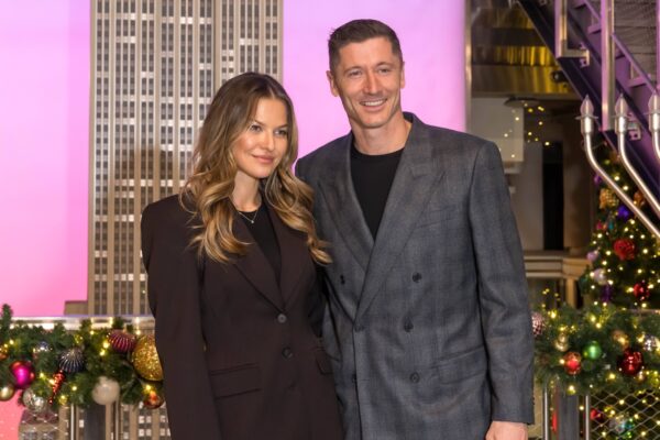 Lewandowscy na Empire State Building. Na tym zdjęciu widzimy uśmiechniętych Roberta i Annę Lewandowskich na Empire State Building w eleganckich strojach. Para pozuje na tle podświetlonej na różowo-fioletowo ściany z wizerunkiem wieżowca, co nadaje ujęciu nowoczesny charakter. Robert ma na sobie szary garnitur w kratę, a Anna ciemny, dopasowany komplet. Tło jest dodatkowo ozdobione świątecznymi dekoracjami, z girlandami i bombkami, co sugeruje, że wizyta odbyła się w okresie przedświątecznym lub świątecznym, uświetniając polskie Święto Niepodległości.