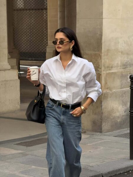 Kobieta spacerująca w miejskim otoczeniu, niosąca kubek kawy. Ubrana jest w trzy absolutnie ponadczasowe elementy: klasyczną, białą, bawełnianą koszulę (lekko oversize), dżinsy w kolorze niebieskiego spranego denimu o luźnym, prostym kroju oraz elegancką, czarną torebkę o sztywnej formie. Stylizację podkreśla czarny, skórzany pasek ze złotą klamrą oraz biżuteria i okrągłe okulary przeciwsłoneczne, co tworzy kwintesencję francuskiego, niewymuszonego stylu.