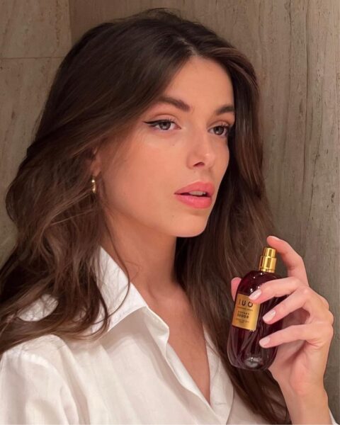 Portret kobiety o długich, brązowych włosach, ubranej w klasyczną, białą koszulę, która trzyma w dłoni elegancki, ciemny flakon perfum. Jej spojrzenie jest intensywne, a makijaż podkreśla naturalne rysy. Flakon o głębokim, rubinowym kolorze i złotej nakrętce sugeruje skomplikowane i luksusowe kompozycje, być może z akordami paczuli i wetywerii, czyli nut zapachowych, które mają być popularne w 2026 roku. 