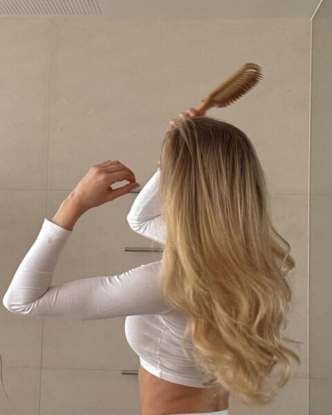 Kobieta o długich, lśniących, jasnoblond włosach czesze się drewnianą szczotką przed lustrem w jasnej łazience. Jej gęste, falujące pasma symbolizują zdrową kondycję. Taki wygląd jest często pożądany, zwłaszcza w kontekście dyskusji na temat ewentualnego osłabienia lub wypadania włosów związanego z szybką utratą wagi. Zdjęcie przedstawia dbałość o mocne i bujne włosy po ozempicu.