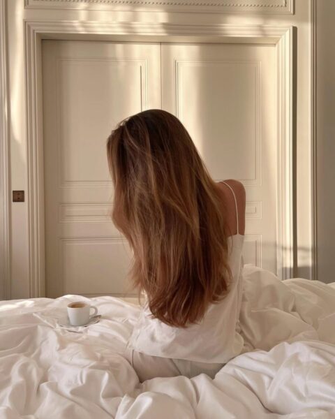 Kadr z tyłu przedstawia kobietę siedzącą na łóżku z kubkiem kawy, w białej bieliźnie nocnej, otoczoną promieniami słońca. Osoba ta ma długie, gęste i zdrowe, brązowo-blond włosy, które spływają na jej plecy, zakrywając twarz. Jest to idealna wizualizacja kondycji włosów, o którą dbają osoby doświadczające ewentualnych problemów związanych z kuracją odchudzającą. Zdjęcie symbolizuje pożądany, zdrowy stan włosów po ozempicu – już po regeneracji.