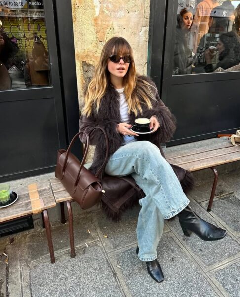 Kobieta siedząca na drewnianej ławce przed kawiarnią, z filiżanką kawy w ręku. Ubrana jest w klasyczne, jasne dżinsy typu "mom jeans" lub proste, luźne spodnie, zestawione z ponadczasową, prostą, białą koszulką (T-shirtem). Całość przełamuje długi, czekoladowy płaszcz ze sztucznego futra lub kożuch, element, który co sezon powraca na jesień i zimę. Stylizację uzupełniają czarne botki z kwadratowym noskiem, dodające nowoczesności, oraz elegancka, skórzana torebka w kolorze czekoladowego brązu – torebka typu tote lub worek, która jest biznesowym i codziennym klasykiem.