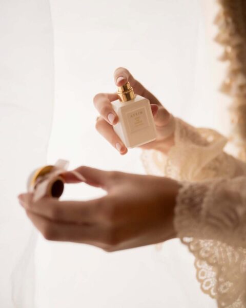 Zbliżenie na dłonie trzymające kremowy flakon perfum AERIN Waterlily Sun ze złotym korkiem, na tle jasnej, tiulowej tkaniny. Kobieta w beżowej koronkowej bluzce przygotowuje się do aplikacji zapachu. Idealne jako miodowe perfumy na jesień 2025.