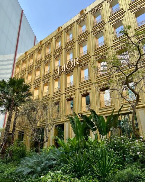 Najbardziej luksusowe sklepy modowe na świecie. Złota fasada wielopiętrowego budynku butiku Dior, zdobiona rzędami klasycznych okien z balustradami, emanuje majestatem i bogactwem. Przed nią rozciąga się bujny, tropikalny ogród z palmami i egzotyczną roślinnością, tworząc zaskakujący kontrast między architekturą a naturą. Centralnie umieszczone, podświetlane logo marki dodaje prestiżu tej unikalnej konstrukcji. Całość tworzy imponującą wizytówkę marki w otoczeniu zieleni.