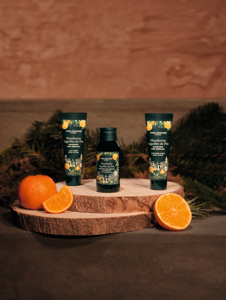 Komplet świątecznych kosmetyków Yves Rocher Mandarine et Aiguilles de Pin ustawiony na drewnianej podstawie, otoczony gałązkami jodły i połówkami soczystej mandarynki. Ujęcie w ciepłym świetle tworzy naturalny, zimowy klimat.