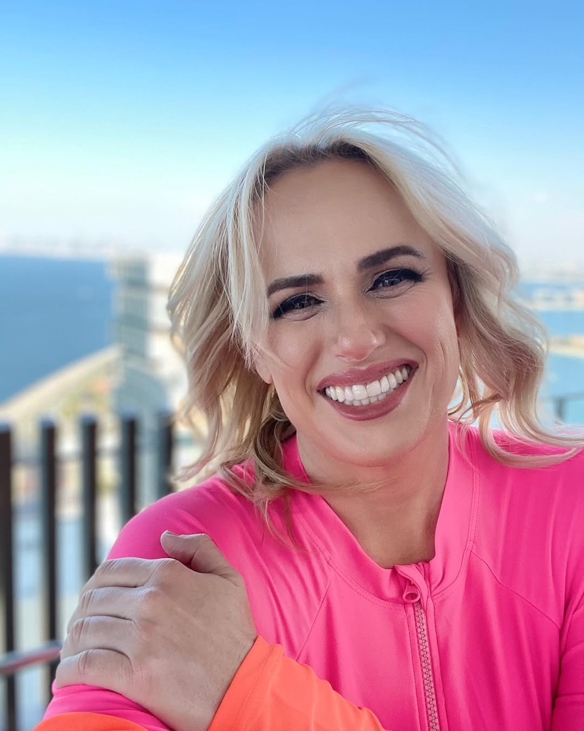 Aktorka Rebel Wilson uśmiecha się szeroko do kamery w jaskrawej, różowej bluzie sportowej, pozując na balkonie z widokiem na morze i miasto w tle. Jej promienny uśmiech z zadbanymi, białymi zębami jest centralnym punktem tego słonecznego ujęcia. Zdrowe zęby są kluczowe dla publicznego wizerunku, co jest szczególnie istotne w świetle doniesień o wpływie terapii odchudzających. Pacjenci stosujący popularne leki na utratę wagi muszą podjąć dodatkowe środki ostrożności, aby ich kuracja nie doprowadziła do problemów stomatologicznych znanych jako ozempicowe zęby.