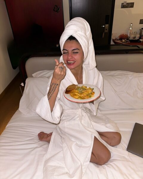 Kobieta w białym, luksusowym szlafroku i ręczniku frotte zawiązanym na głowie (po relaksującej kąpieli) siedzi w po turecku na białym łóżku hotelowym lub sypialnianym. Z szerokim, radosnym uśmiechem i widelcem w dłoni delektuje się ciepłym posiłkiem.