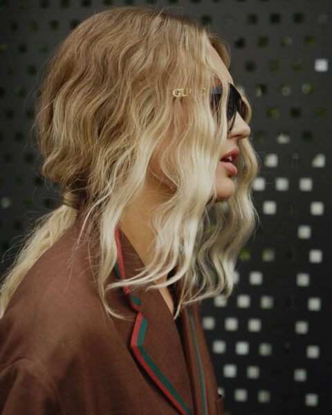 Profil kobiety z falowanymi, blond włosami, częściowo spiętymi w luźny kucyk. Kobieta ma duże okulary przeciwsłoneczne i brązową marynarkę z zielono-czerwonymi detalami.
