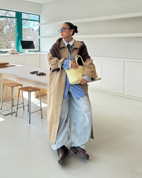 Kobieta w stylowym, długim beżowo-brązowym trenczu, luźnych dżinsach i biało-niebieskiej koszuli w paski stoi w jasnym, nowoczesnym wnętrzu. Trzyma w ręku plecioną, żółtą torebkę i ma na sobie modne, niebieskie okulary przeciwsłoneczne. W tle znajduje się drewniany stół i białe półki.