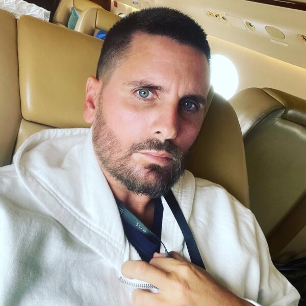 Scott Disick robi selfie wewnątrz prywatnego odrzutowca, siedząc w jasnobeżowym fotelu i mając na sobie białą bluzę z kapturem. Jego intensywnie niebieskie oczy kontrastują z ciemną, schludną brodą. Wyraźnie szczuplejszy wygląd celebryty, widoczny na tym zbliżeniu, nawiązuje do szeroko komentowanych spekulacji o stosowaniu Ozempicu w Hollywood. Ta szybka transformacja Disicka wzbudzała duże zainteresowanie mediów.