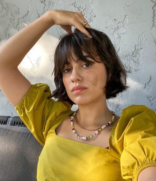 Zdjęcie przedstawia młodą kobietę o ciemnych, krótkich włosach Chin-length bob z grzywką, patrzącą prosto w obiektyw. Ma na sobie musztardowożółtą bluzkę z bufiastymi rękawami i kwadratowym dekoltem. Nosi również naszyjnik z różnokolorowymi koralikami i perłami oraz złote pierścionki. Jedną rękę trzyma na czubku głowy. Tło stanowi jasna, teksturowana ściana.