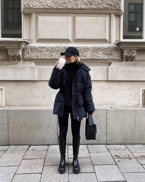 Ta monochromatyczna, czarna stylizacja stanowi idealne nawiązanie do trendu quiet luxury, czyli dyskretnego luksusu. Kobieta ubrana jest w czarny golf, spodnie i efektowne, wysokie botki na grubej podeszwie. Centralnym punktem jest czarna puchówka z sieciówki o regulowanej talii, która łączy modę i funkcjonalność. Ta kurtka, w połączeniu z czapką bejsbolową, to obowiązkowy element, który będzie królował w kolekcjach na zimę 2025.