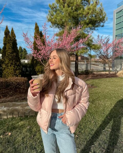 Młoda kobieta w wiosenno-zimowej stylizacji pozuje z kawą na tle różowo kwitnących drzew. Ubrana jest w modną, krótką, jasnoróżową kurtkę puchową, biały crop top i jasne jeansy z wysokim stanem. Ten sportowo-elegancki zestaw świetnie oddaje ducha mody ulicznej, w której puchówki z sieciówki są kluczowym elementem. Kurtka tego typu to idealny fason, który z pewnością zobaczymy w kolekcjach, gdy nadejdzie zima 2025.