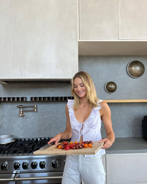 Uśmiechnięta, młoda kobieta w eleganckiej, białej bluzce z falbankami i białych spodniach, stoi w nowoczesnej, minimalistycznej kuchni. Trzyma przed sobą drewnianą deskę do krojenia z garścią kolorowych, świeżo pokrojonych pomidorów koktajlowych. W tle widać luksusowy, profesjonalny piec kuchenny oraz stylowe, betonowe ściany i designerskie akcenty, co sugeruje kulinarną pasję w wnętrzu godnym katalogu. To scena idealna, by stworzyć dania, których zapach wypełni dom jak najlepsza restauracja śródziemnomorska.