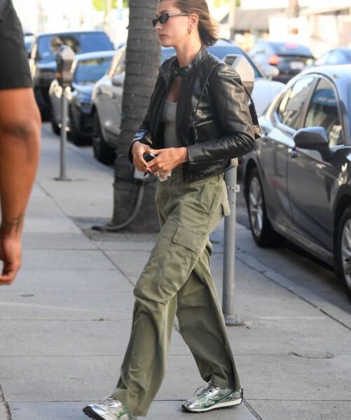 Hailey Bieber idzie po chodniku, ubrana w czarną skórzaną kurtkę typu biker, szary crop top, szerokie spodnie cargo w kolorze oliwkowej zieleni oraz sportowe buty w metalicznym odcieniu srebra z zielonymi akcentami. Ma włosy spięte w kok i ciemne okulary przeciwsłoneczne. W tle widać zaparkowane samochody i drzewa.