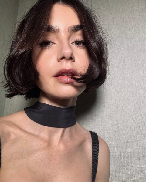 Zdjęcie przedstawia młodą kobietę Lily Collins o ciemnobrązowych, gładkich włosach, ściętych w elegancki bob do linii szczęki. Kobieta ma wyraziste brwi i patrzy prosto w obiektyw. Ma na sobie czarny top na ramiączkach i czarną, szeroką opaskę (choker) na szyi. Tło jest szare i teksturowane.