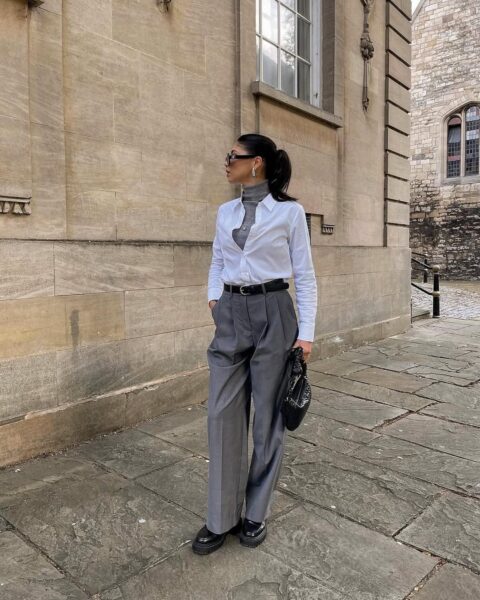 Kobieta ubrana w szerokie, szare spodnie z wysokim stanem, szary golf i rozpiętą białą koszulę stoi na chodniku z kamiennych płyt przed budynkiem z jasnego kamienia. Trzyma w ręku małą, czarną torebkę i ma na sobie czarne buty na grubej podeszwie. Nosi okulary przeciwsłoneczne i duże kolczyki koła.