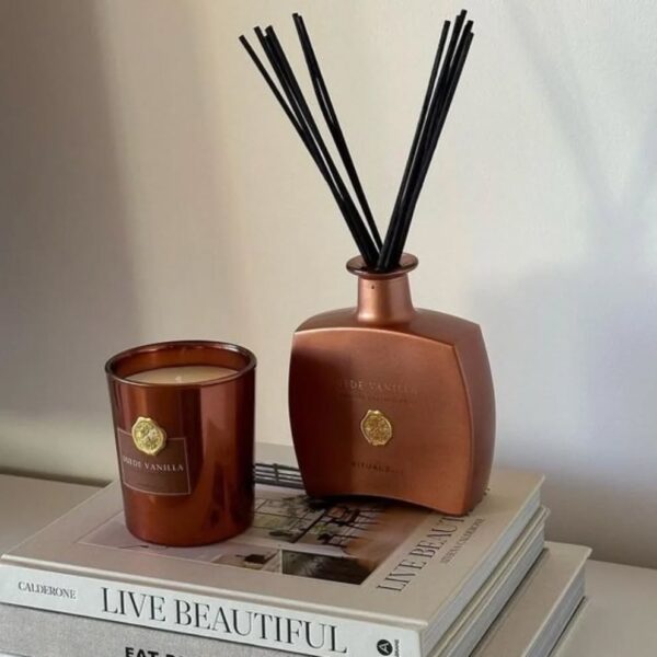 Luksusowa aranżacja zapachowa na stercie eleganckich książek. Na pierwszym planie widoczny jest zestaw do aromaterapii w głębokim, miedzianym lub brązowym odcieniu: gruba, szklana świeca zapachowa oraz dyfuzor patyczkowy w wyrafinowanej, kanciastej butelce, oba opatrzone złotym logo. Etykieta świecy głosi "SUEDE VANILLA". Całość emanuje spokojem i elegancją, idealnie pasując do wnętrza pachnącego jak ekskluzywny hotel lub orientalny ogród.