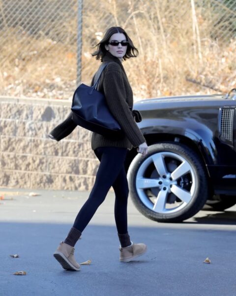 Kendall Jenner idzie po chodniku w swetrze w kolorze khaki lub ciemnozielonym, czarnych legginsach i krótkich beżowych botkach UGG. Ma ciemne, sięgające ramion włosy, ciemne okulary przeciwsłoneczne i niesie dużą czarną torbę na ramię. W tle widać czarnego SUV-a i suchą roślinność.