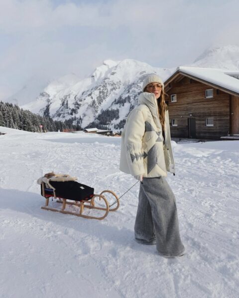 Kobieta w górskiej scenerii ciągnie sanki z dzieckiem, ubrana w modną, białą puchówkę z geometrycznym wzorem. Ta puchówka z sieciówki w stylu après-ski doskonale łączy funkcjonalność z zimowym szykiem. To przykład na to, jak puchówki z sieciówki na zimę 2025 będą stawiać na wyraziste wzory i wysoką jakość.