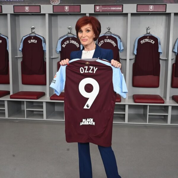Sharon Osbourne, znana z otwartego stosowania leku Ozempic i wynikającej z niego znacznej utraty wagi, stoi w szatni klubu West Ham United. Ubrana w elegancki, granatowy garnitur, trzyma dwiema rękami klubową koszulkę piłkarską z numerem 9, nazwiskiem "OZZY" i napisem "BLACK SABBATH". Jej wyraźnie szczuplejsza sylwetka i charakterystyczna fryzura są widoczne na tle szafek z koszulkami rockowymi.