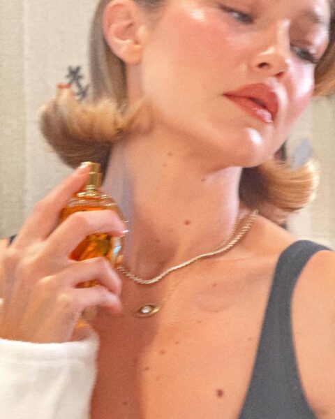 Kobieta o jasnej karnacji, z włosami ułożonymi w delikatne fale, spryskuje swoją szyję miodowymi perfumami z eleganckiego, bursztynowego flakonu. Ma na sobie cienki, czarny top i naszyjnik z błyszczącymi kamieniami i małą zawieszką. Scena jest dobrze oświetlona, z ciepłym, rozproszonym światłem.