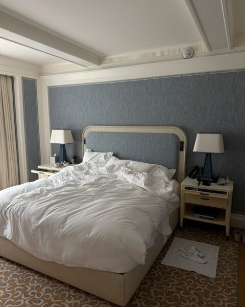 Wnętrze luksusowej sypialni o klasycznym designie. Centralnym elementem jest duże łóżko z wysokim, kremowym zagłówkiem i mnóstwem śnieżnobiałej, pogniecionej pościeli, która sprawia wrażenie niesamowicie wygodnej. Ściana za łóżkiem jest pokryta elegancką, błękitno-szarą tapetą z delikatną fakturą, obramowaną sztukaterią. Po obu stronach stoją beżowe szafki nocne z designerskimi lampami na niebieskich podstawach. Całość uzupełnia wykładzina w klasyczny, geometryczny wzór w odcieniach złota i beżu. To idealny widok, który przywodzi na myśl sypialnię w pięciogwiazdkowym hotelu lub dom pachnący jak najbardziej ekskluzywne miejsce na Riwierze Francuskiej.