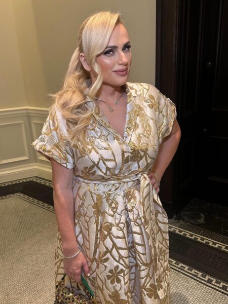 Zdjęcie przedstawia aktorkę Rebel Wilson, stojącą w eleganckim wnętrzu z panelami ściennymi i mozaikową podłogą. Ma na sobie elegancką sukienkę kopertową z krótkimi rękawami, wykonaną z bogatej tkaniny w kolorze kremowym lub złotym z metalicznym, złotym, żakardowym wzorem kwiatowym/liściastym. Sukienka jest przewiązana w talii pasem z tej samej tkaniny. Wilson pozuje z pewnym uśmiechem, trzymając rękę na biodrze. Jej blond włosy są częściowo upięte z długą, pofalowaną resztą spływającą na ramię, a na szyi ma delikatny naszyjnik.
