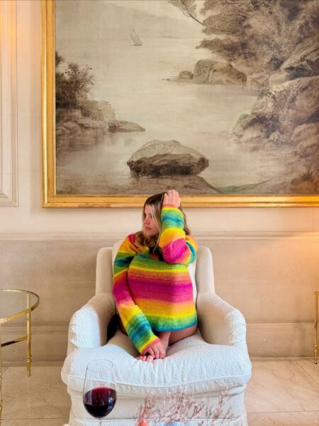 Ujęcie kontrastu i luksusu. Kobieta siedzi z podciągniętymi kolanami w wygodnym, kremowym, tapicerowanym fotelu, umieszczonym w klasycznie urządzonym, eleganckim salonie. Ma na sobie miękki, oversizowy sweter w jaskrawe, tęczowe pasy, który stanowi wyrazisty, nowoczesny akcent na tle tradycyjnego wnętrza. Za nią wisi duży, zabytkowy obraz w masywnej złotej ramie, przedstawiający klasyczny, malowniczy pejzaż. Na stoliku w pobliżu stoi kieliszek czerwonego wina. Cała scena łączy luksus i komfort z artystyczną ekstrawagancją, tworząc atmosferę prywatnej galerii w Paryżu czy salonu we włoskiej willi.