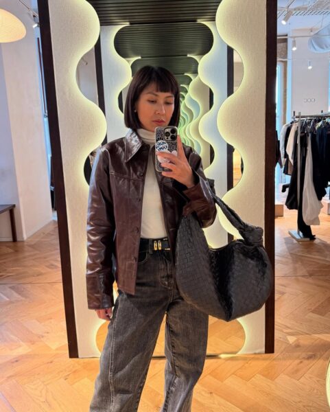 Zdjęcie przedstawia kobietę robiącą sobie selfie w lustrze w sklepie. Ma ciemne, proste włosy w stylu bob do linii szczęki z krótką grzywką. Jest ubrana w biały golf, brązową skórzaną kurtkę i ciemne, luźne dżinsy. Przewieszoną przez ramię trzyma dużą, plecioną, czarną torbę. Kobieta trzyma telefon w etui z napisem w dłoni, zasłaniając część twarzy. Tło stanowią lustra o falistych, podświetlonych ramach i drewniana podłoga w jodełkę.