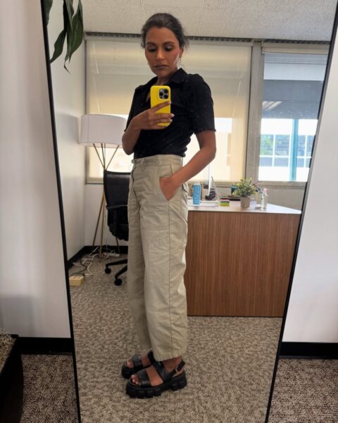 Zdjęcie przedstawia Mindy Kaling, która robi sobie lustrzane selfie w biurze. Ma na sobie stylowy, casualowy zestaw: czarną, dopasowaną bluzkę polo z krótkim rękawem oraz luźne, beżowe spodnie typu cargo z podwyższonym stanem i kieszeniami, z jedną ręką w kieszeni. Stylizację dopełniają masywne, czarne sandały na grubej platformie. Trzyma w dłoni telefon w jasnym, żółtym etui, który zakrywa część jej twarzy. W tle widać przestrzeń biurową z biurkiem, krzesłem biurowym i dużymi oknami.