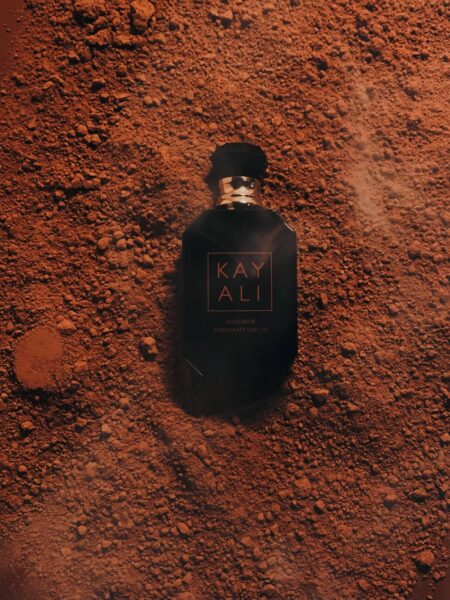 Kayali OUDGASM Chocolate Oud || 11, czarny flakon perfum leżący na brązowym, pylistym proszku przypominającym kakao lub ziemię. Doskonałe czekoladowe perfumy z nutą oudu.