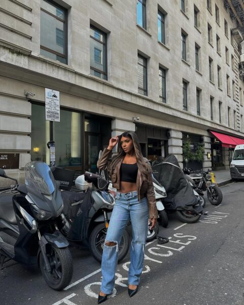 Kobieta o długich, brązowych włosach pozuje na ulicy obok rzędu zaparkowanych skuterów i motocykli. Ma na sobie krótki czarny top, brązową skórzaną kurtkę typu crop oraz luźne, jasne jeansy z dziurami na kolanach. Nosi czarne, spiczaste szpilki i trzyma ciemne okulary przeciwsłoneczne. W tle widać wysoką, kamienną, wielopiętrową kamienicę.