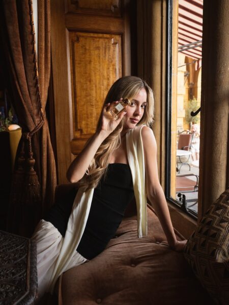 Elegancka kobieta w czarno-białej sukience siedzi przy oknie, trzymając w dłoni prostokątny flakon perfum. Tło stanowią luksusowe, drewniane elementy wnętrza i brązowe zasłony.