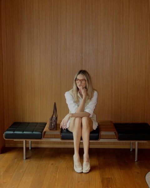 Stylowa, młoda kobieta w eleganckiej, casualowej stylizacji – biała koszula, jasna kamizelka i beżowe mokasyny – siedzi na designerskiej, modernistycznej ławce z czarną, skórzaną tapicerką. Ma na sobie modne, duże okulary i wydaje się być zamyślona, opierając brodę na dłoni. Tłem jest ściana wyłożona ciepłymi, drewnianymi panelami w pionowe paski. Całość emanuje spokojem i wyrafinowaniem, przypominając poczekalnię luksusowego butiku w Mediolanie lub gabinet designera z połowy XX wieku. Styl, który sprawia, że czujesz się, jakbyś znalazł się w najbardziej eleganckim miejscu na świecie.